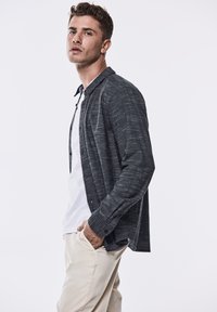 Tmavě šedá košile na knoflíky s texturovaným vzorem, nošená přes bílý tričko, doplněná světlými kalhotami. Model stojí bokem.