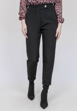 Femme portant un pantalon noir taille haute à jambes fuselées avec ourlets retroussés et un chemisier à manches longues à motifs, assortis à des bottines noires à talons.