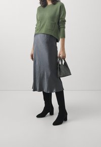 Maglione verde a maglia con due tasche frontali, abbinato a una gonna midi in satin grigia. Stivali neri al ginocchio e una borsa texturizzata completano l'outfit.