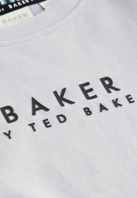Balta kokvilnas T-kreklā ir tekstūru melna druka ar uzrakstu "BAKER" un "by TED BAKER". Apaļš kakls ar redzamu šuvju detaļu.