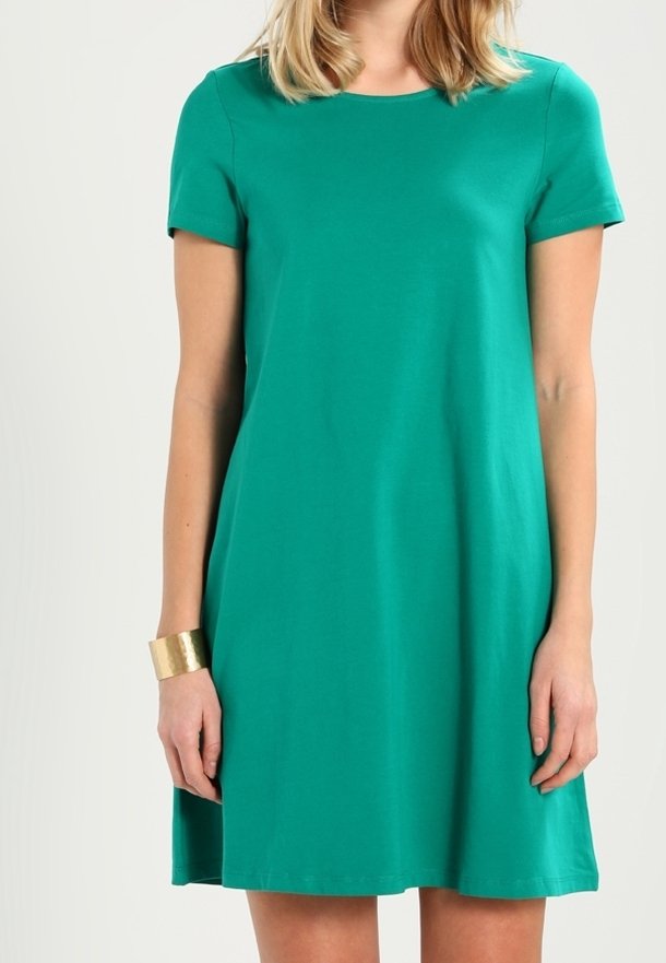 Vrouw in een groene jurk met korte mouwen tot op de knie en een gouden armband om haar linkerpols.