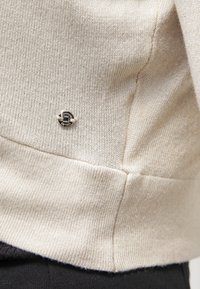 Beige pletený svetr s ribkovanou texturou, s malým kulatým kovovým logem poblíž lemu a jemnými detaily v šití.