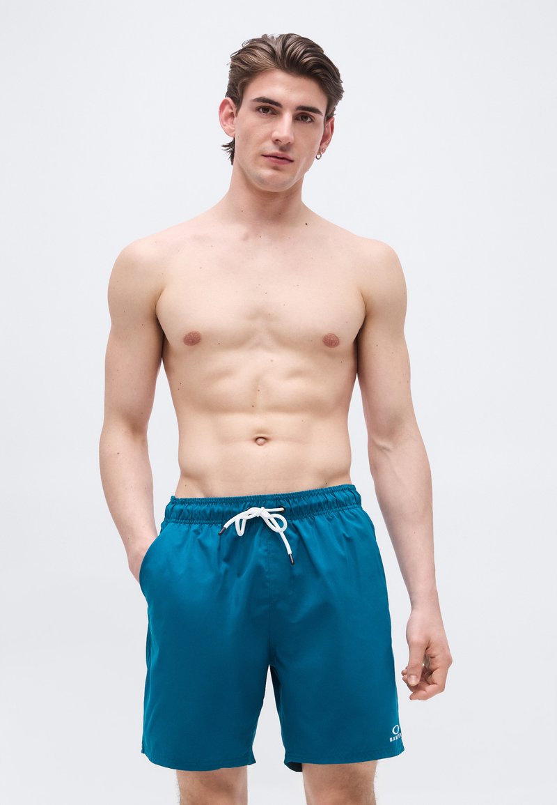 Άνδρας με κοντά καστανά μαλλιά στέκεται χωρίς πουκάμισο φορώντας μπλε μαγιό με λευκό κορδόνι, έχοντας το ένα χέρι στην τσέπη σε απλό φόντο.