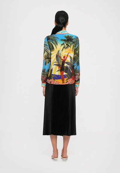 Blouse à manches longues colorée avec un motif tropical, associée à une jupe noire plissée. La blouse présente un décolleté vibrant avec un motif.