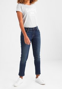 Šedé tričko s krátkým rukávem s logem "GANT", spojené s tmavě modrými džíny slim fit a bílými teniskami. Neformální, padnoucí styl.