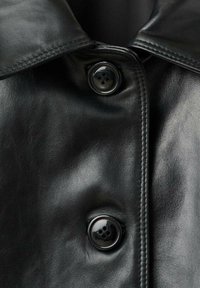 Czarna skórzana kurtka z dwoma dużymi, błyszczącymi czarnymi guzikami i szczegółowym wykończeniem w okolicach kołnierza i guzików.