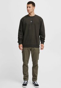 Dunkelolivgrüner Sweatshirt mit Rundhalsausschnitt und gerippten Bündchen, kombiniert mit olivfarbenen Cargo-Hosen und schwarzen Turnschuhen. Kleines Logopatch auf der Brust.