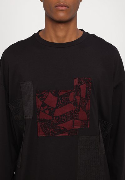 Chemise noire à manches longues avec un patch maroon texturé présentant des motifs complexes, rehaussée de compléments en tissu noir.