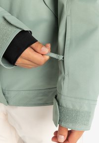 Grøn jakke med en glat, struktureret overflade; har justerbar manschet med velcro og en trækstrop; quiltet detalje ved den nederste søm.