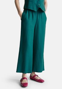 Pantaloni larghi color turchese con cintura elastica indossati con un gilet coordinato e scarpe Mary Jane rosa brillante con cinturini a fibbia.