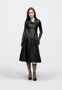 Melna ādas kleita ar pieguļošu augšdaļu, priekšā rāvējslēdzēju un garām piedurknēm. Svārki izvērsti, garums līdz ceļiem. Valdzēta ar cimdiem un papēžu kurpēm ar potītes siksnu.