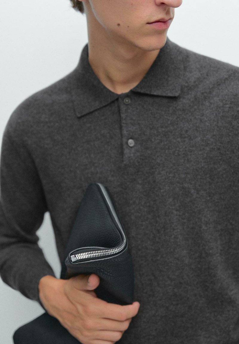 Donkergrijs gebreid sweater met een kraag, twee knopen en een gladde textuur. Het model houdt een zwarte clutch met rits en een gestructureerde afwerking vast.