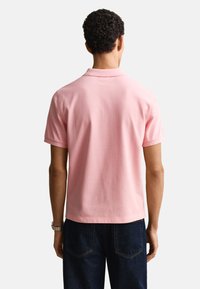 Muž s krátkymi kučeravými vlasmi, ktorý má na sebe svetloružové polo tričko s krátkym rukávom a tmavomodré džínsy, zobrazený zozadu na bielom pozadí.