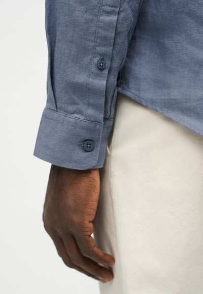 Σκοτεινόχρωμο χέρι που ακουμπά δίπλα σε ανοιχτό μπεζ παντελόνι, φοράει μπλε πουκάμισο με μακριά μανίκια και κουμπωμένα μανικόφυλλα.