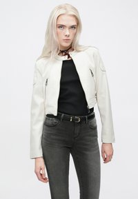 Λευκή ψεύτικη δερμάτινη cropped ζακέτα με φερμουάρ και ομαλή υφή, φορεμένη πάνω από μαύρο μπλουζάκι και συνδυασμένη με γκρι τζιν και μαύρη ζώνη.