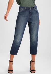 Jeans de denim azul con una cintura alta y un largo recortado, que presentan parches en las rodillas, diseño de cinco bolsillos y un sutil desvanecimiento.