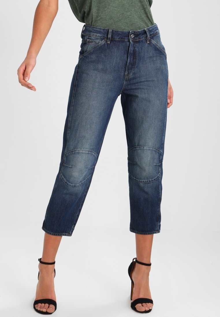 Jeans de denim azul con una cintura alta y un largo recortado, que presentan parches en las rodillas, diseño de cinco bolsillos y un sutil desvanecimiento.