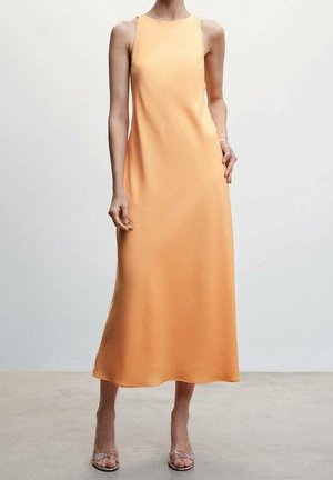 Femme portant une robe sans manches couleur pêche, longue jusqu'aux chevilles, avec des sandales à talons transparents, debout sur un sol uni.