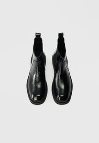 Un par de botas Chelsea de cuero negro brillante con paneles elásticos en los laterales y tiradores, vistas desde arriba sobre un fondo liso.