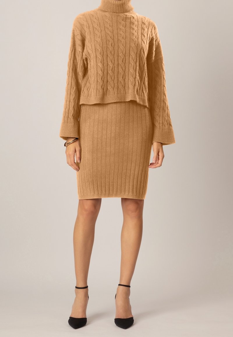 Femme portant un pull à col roulé en maille torsadée couleur camel et une jupe crayon côtelée, assortis à des talons à bride noire autour de la cheville et un bracelet.