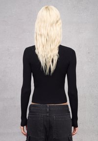 Top de manga larga negro acanalado con diseño ajustado, combinado con pantalones de mezclilla oscuros de talle alto que tienen bolsillos laterales. Cabello rubio ondulado visible.