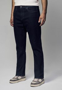 Jeans de mezclilla oscura con un diseño de pierna recta, estilo clásico de cinco bolsillos y costuras en contraste. Combinados con zapatillas grises y blancas.