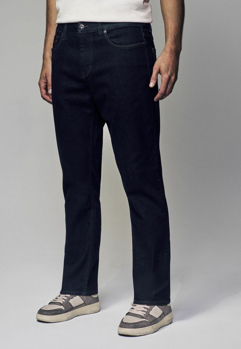 Jeans de mezclilla oscura con un diseño de pierna recta, estilo clásico de cinco bolsillos y costuras en contraste. Combinados con zapatillas grises y blancas.
