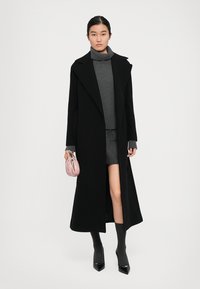Woman wearing a long black coat over a gray turtleneck sweater and skirt, holding a small pink handbag, standing against a white background.
