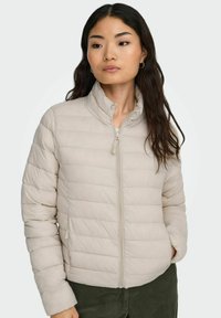 Gaiši beigu krāsas džinsis ar stāvjo apkakli, priekšējo rāvējslēdzēju un sānu kabatām; tam ir horizontālas šuves un gluda tekstūra.