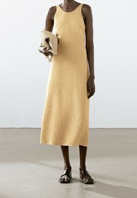 Leinenkleid in Gelb mit Rundhalsausschnitt und Midi-Länge, kombiniert mit einer beigen geflochtenen Tasche. Das Modell trägt schwarze flache Sandalen.
