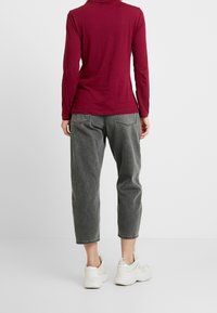 Bordeaux top met lange mouwen, gecombineerd met grijze cropped broek. De stof lijkt zacht, de broek heeft zakken en de look wordt afgerond met witte sneakers.