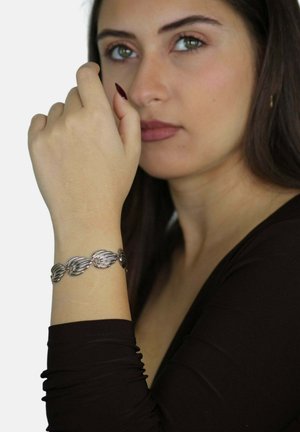 Bracciale d'argento con un design a foglia, caratterizzato da forme lisce e ondulate. Indossato su un polso, con un top a maniche lunghe scuro visibile.