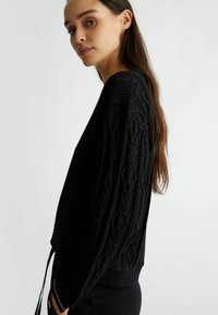 Maglione nero con maniche a treccia testurizzate, orlo a costine e lacci laterali. Realizzato in tessuto morbido con una leggera finitura lucida.