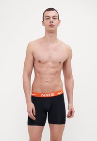 Fekete boxer alsónadrág, rugalmas narancssárga derékszíjjal, amelyen a Nike logó található. Az anyag simának és testhezállónak tűnik. A modell semleges testtartásban van.