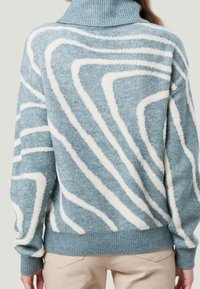 Frau in einem blauen Rollkragensweater mit weißen abstrakten Wirbelmustern, kombiniert mit hellbeigen Hosen, von hinten betrachtet.