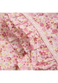 Blommönstrat tyg med små rosa och gula blommor. Dekorativa volanger och spetsdetaljer längs ena sidan ger textur och detaljer.