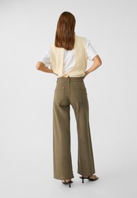 Lichtbeige gebreide vest over een wit T-shirt, gecombineerd met olijfgroene wijde broek. Zwarte hoge hak sandalen maken de look compleet.