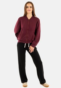 Felpa con cappuccio color bordeaux con cerniera e tasche laterali, abbinata a pantaloni neri a gamba larga con vita con coulisse e scarpe beige. Tessuto liscio.