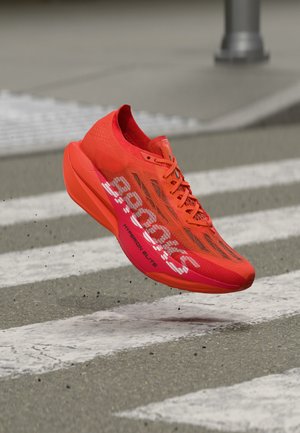 Svetlo oranžová bežecká obuv so šnúrkami, s brandingom "Hoka One One" a "Hyperion Elite", vznášajúca sa nad priechodom pre chodcov.