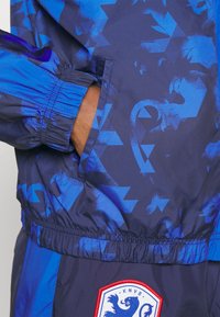 Blå jacka med dragkedja och geometriskt mönster, elastiska muddar och midjeband. Har en KNVB-logotyp i rött på en vit lapp.