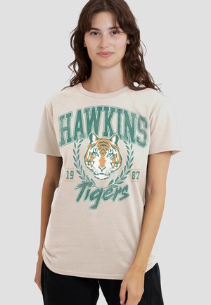 Beige cotton t-shirt featuring a green and orange tiger graphic, "HAWKINS" text above, and "Tigers" below, with laurel accents and a round neckline.