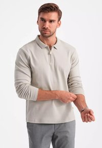 Hellgraues Langarm-Polo mit halbem Reißverschluss am Kragen. Weicher Stoff, entspannte Passform und umgeschlagene Bündchen. Wird mit grauen Hosen für einen abgestimmten Look getragen.