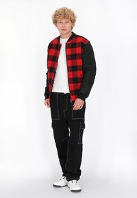 Rood-zwart geruit jack met zwarte mouwen, gedragen over een wit shirt, gecombineerd met zwarte cargobroek en witte sneakers.