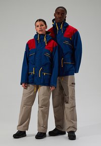 Blaue und rote wasserfeste Jacken mit gelben Akzenten, kombiniert mit beigen Hosen, die über mehrere Taschen und verstellbare Kapuzen verfügen.