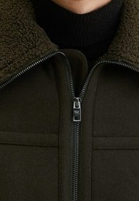 Giacca verde scuro con colletto in fleece e zip frontale, dalla texture liscia e dettagli con zip a contrasto.