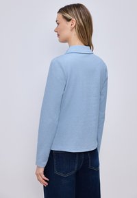 Femme aux cheveux longs et lisses châtain clair, portant un haut bleu clair à manches longues et un jean bleu foncé, de profil sur un fond uni.