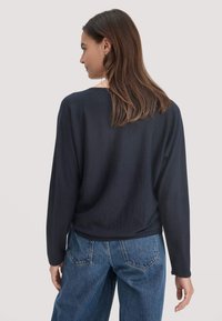 Frau mit braunen Haaren, die ein navyblaues Langarmoberteil und eine blaue Jeans trägt, von hinten vor einem schlichten, hellen Hintergrund gesehen.