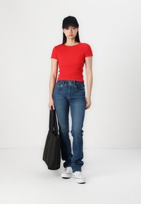 Haut court à manches courtes rouge côtelé, jeans bleus, casquette noire et baskets blanches. Le mannequin tient un sac fourre-tout noir, se tenant devant un fond blanc.