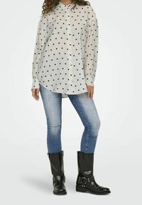 White button-up shirt with black polka dots, long sleeves, and a curved hem, paired with blue jeans and black knee-high boots featuring straps.