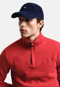 Roter gestrickter Pullover mit Reißverschlusskragen, kombiniert mit einer marineblauen Cordmütze. Die Mütze hat ein gesticktes Logo auf der Vorderseite.